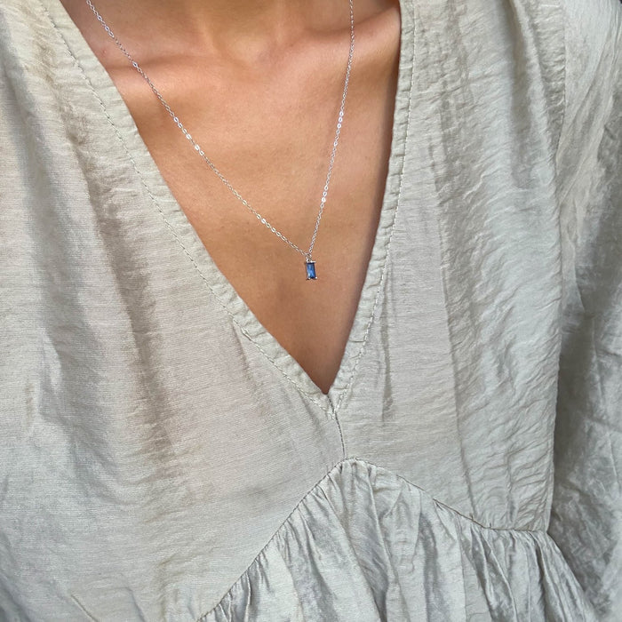 A person wears a textured light beige V-neck blouse paired with the LLUME Jewelry Taylor Necklace Silver, featuring a small sapphire blue charm. The focus is on the blue pendant necklace and neckline.