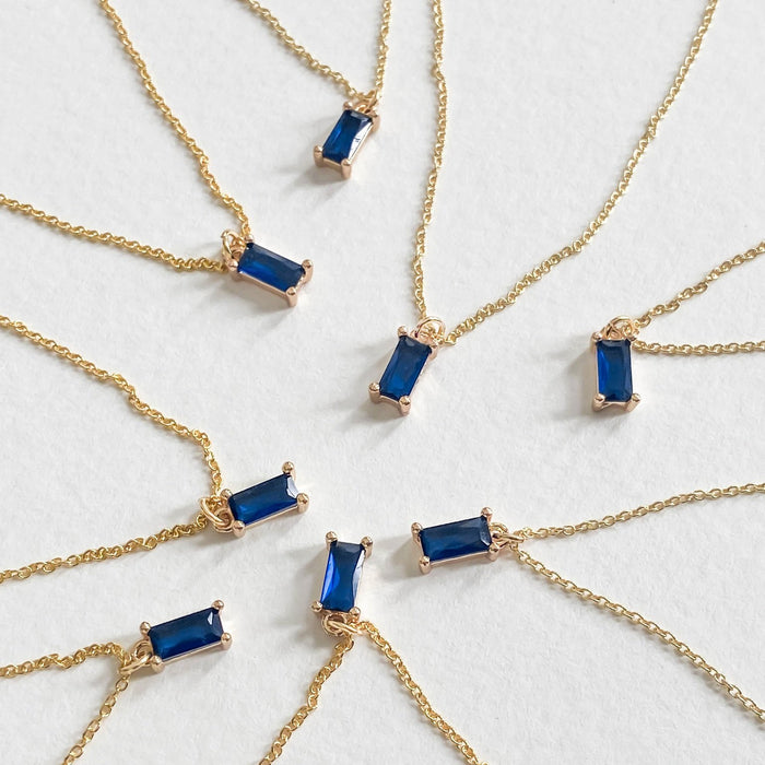 Several Taylor Necklaces by LLUME Jewelry, each featuring a sapphire blue pendant set in a gold-plated frame, are arranged in a circle and displayed on a white background.