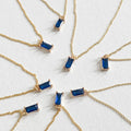 Several Taylor Necklaces by LLUME Jewelry, each featuring a sapphire blue pendant set in a gold-plated frame, are arranged in a circle and displayed on a white background.