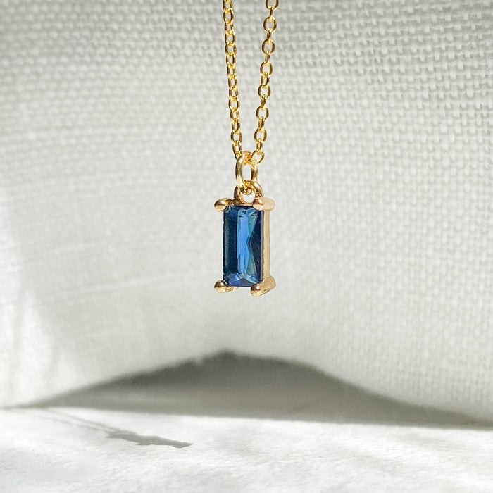 The Taylor Necklace by LLUME Jewelry features a gold plated chain with a rectangular sapphire blue gemstone pendant, shown on textured white fabric under soft lighting—a dainty charm perfect for any occasion.