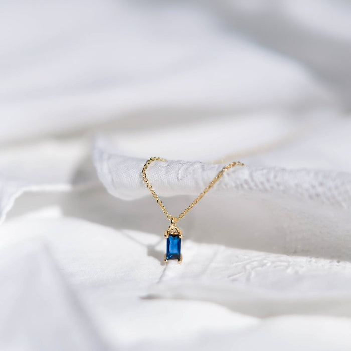 The Taylor Necklace by LLUME Jewelry features a delicate gold-plated chain with a rectangular sapphire-blue pendant, shown resting on soft, subtly textured white fabric.
