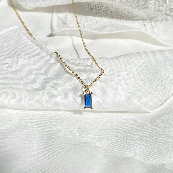 The Taylor Necklace by LLUME Jewelry, featuring a rectangular sapphire blue gemstone pendant with gold plating, is displayed on white textured fabric.