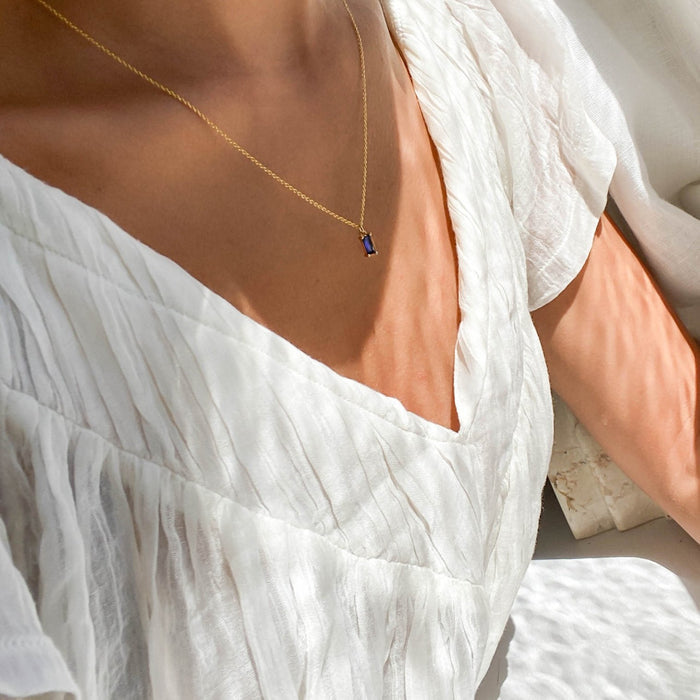 A person wears a white, textured v-neck dress and the LLUME Jewelry Taylor Necklace, a gold plated piece with a small sapphire blue pendant. Soft natural light highlights both the fabric and the necklace.