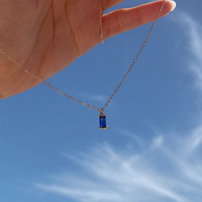 A hand holds the Taylor Necklace by LLUME Jewelry, featuring a delicate blue gemstone pendant, against a bright sky with wispy white clouds.