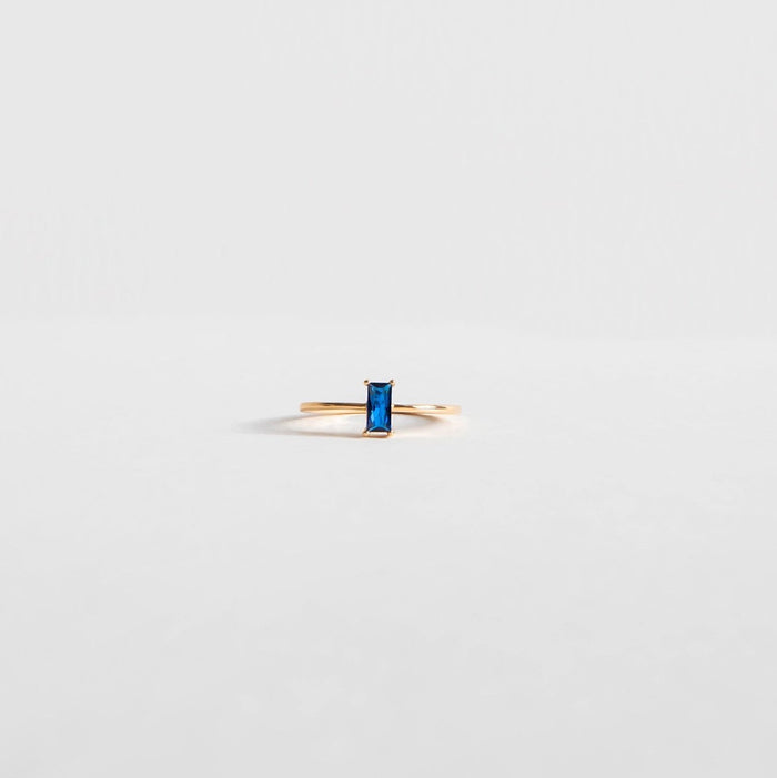 The Taylor Ring by LLUME Jewelry features a 14k gold-filled band with a rectangular blue sapphire, displayed on a plain light gray background.