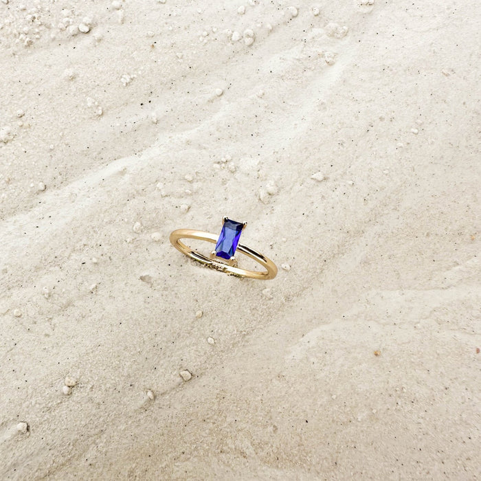 The Taylor Ring by LLUME Jewelry features a rectangular blue sapphire set in 14k gold-filled band, resting on light sand with small pebbles—ideal for stacking or wearing alone.