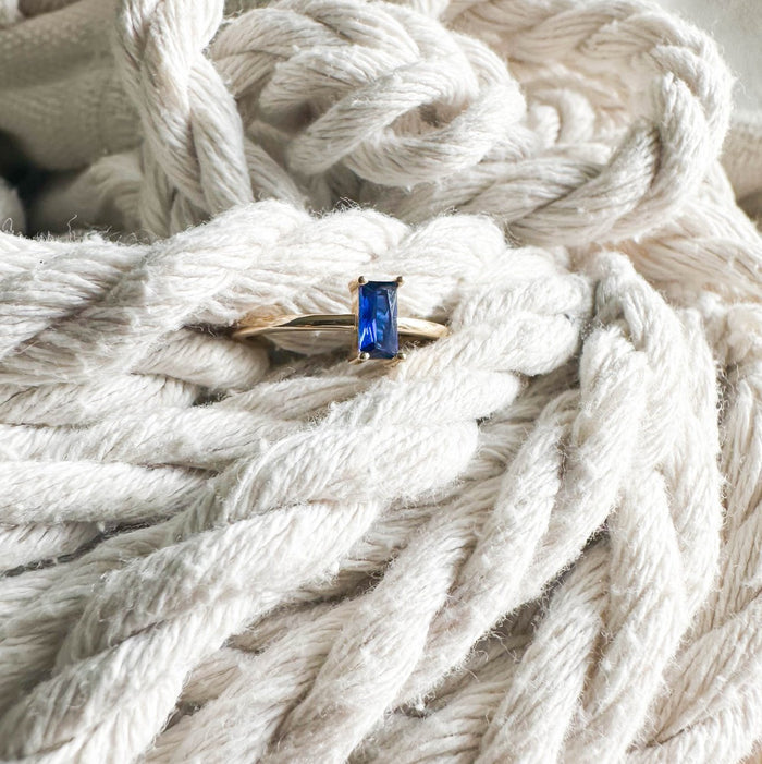 The Taylor Ring by LLUME Jewelry features a rectangular blue sapphire set in 14k gold-filled metal, displayed on thick, white twisted rope.