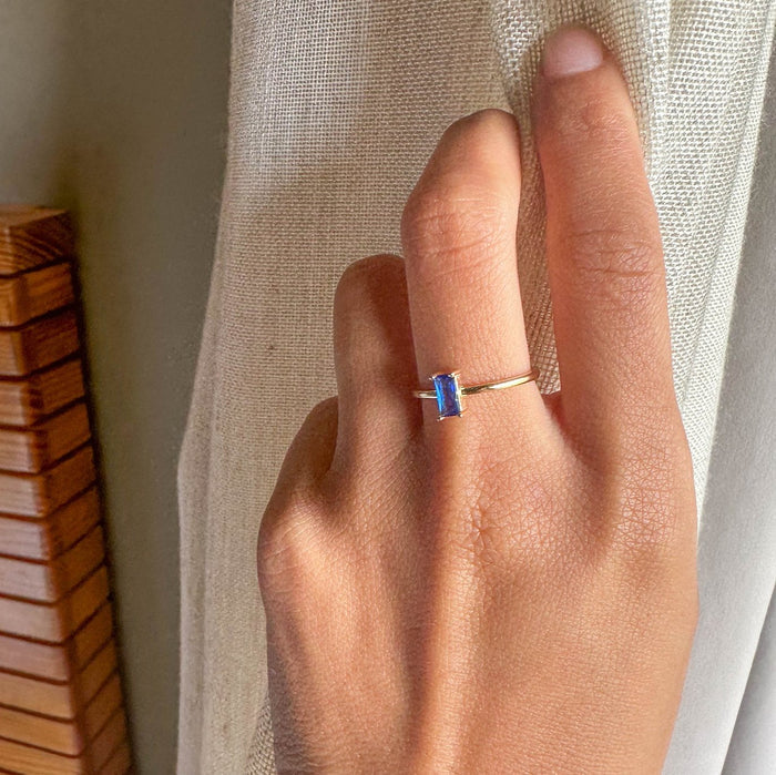 A hand wearing LLUME Jewelry's Taylor Ring, a 14k gold-filled piece with a rectangular blue sapphire, rests on a light curtain with wooden slats in the background.