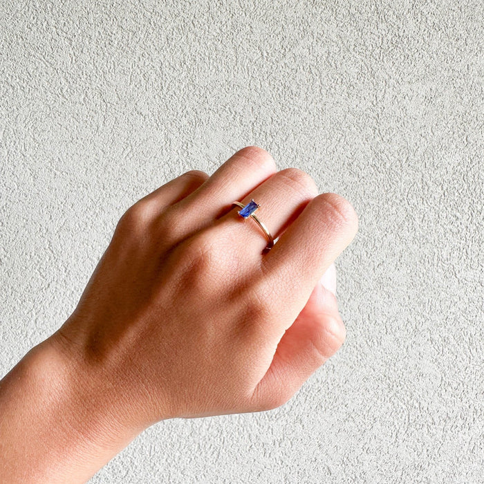 A light-skinned hand wears the Taylor Ring by LLUME Jewelry, a delicate 14k gold-filled stacking ring with a small rectangular blue sapphire, set against a textured light gray background.