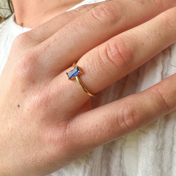 A hand wears the Taylor Ring by LLUME Jewelry—a 14k gold-filled ring with a rectangular blue sapphire—resting on light-textured fabric; ideal for fans of stacking rings.