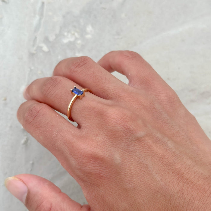 A hand wears the LLUME Jewelry Taylor Ring, a 14k gold-filled design with a rectangular blue sapphire, set against a light textured background.