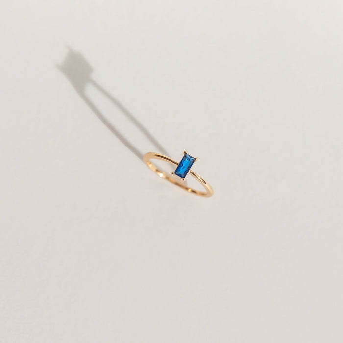 The Taylor Ring by LLUME Jewelry features a rectangular blue gemstone set in delicate 14k gold fill. Shown on a light surface with a soft left shadow, it’s ideal for stacking or wearing alone.