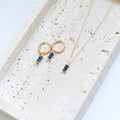 The Taylor Set by LLUME Jewelry, featuring gold hoop earrings with rectangular blue stones and a matching blue pendant necklace, is displayed as a chic set on a textured beige tray.
