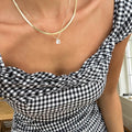 A person wearing a black and white gingham dress with a ruffled neckline is shown from the shoulders down, layering LLUME Jewelry's The Classic Set (retired) while light from a nearby window is visible.