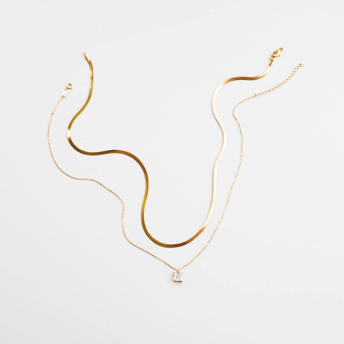 Two delicate gold necklaces from LLUME Jewelry’s retired The Classic Set rest on a white surface. One is a smooth, wavy flat chain; the other features a thin chain with a small square pendant.
