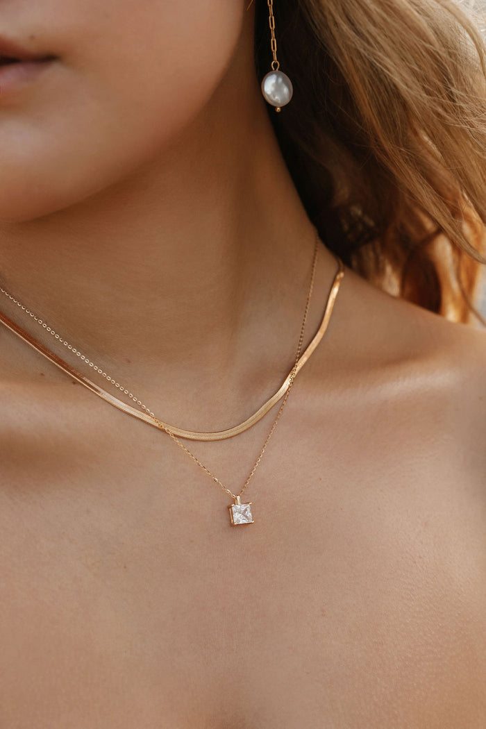 A close-up of a woman’s neck wearing The Classic Set by LLUME Jewelry, featuring a gold herringbone chain, a pendant necklace with a square charm, and a single dangling pearl earring.