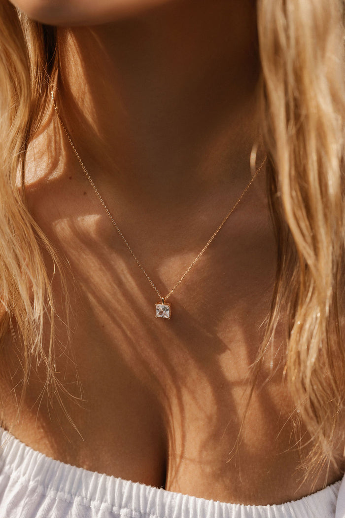 A close-up of a woman wearing LLUME Jewelry’s The One Necklace—a gold plated piece with a square pendant and clear gem charm—against her bare skin, wavy blonde hair, and a textured white top.
