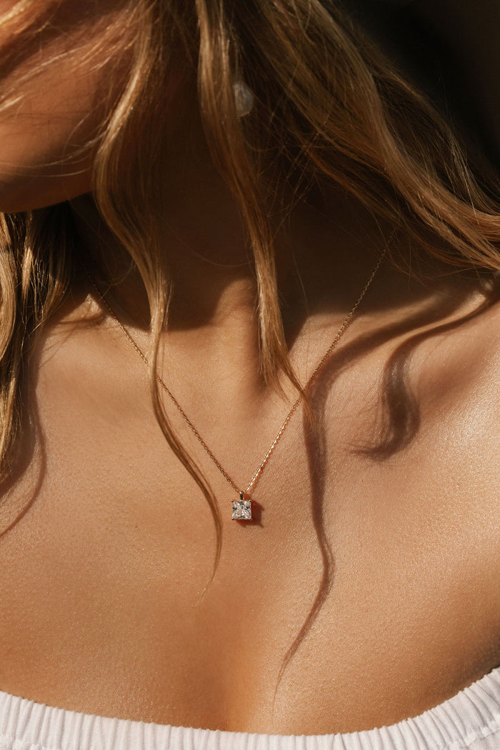 Close-up of a woman's neck and collarbone, with wavy hair, wearing LLUME Jewelry's The One Necklace—a gold plated piece with a square-cut clear gem pendant—styled with a white, off-the-shoulder top.