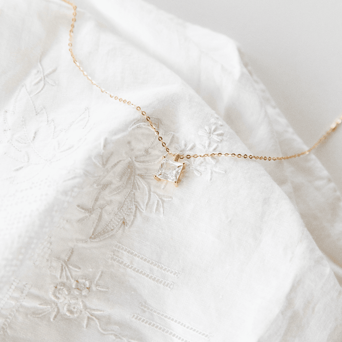 The One Necklace by LLUME Jewelry: a delicate gold vermeil piece with a square clear gemstone pendant, showcased on white embroidered fabric.