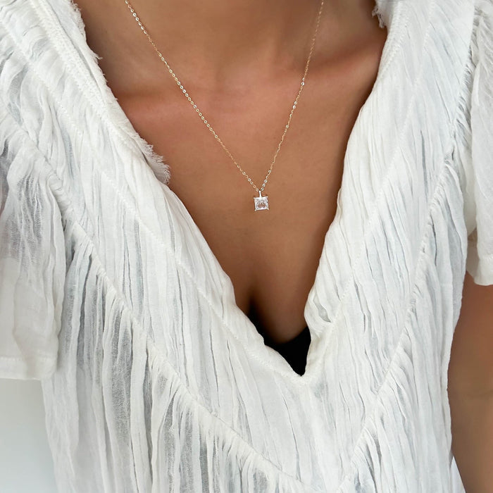 A person wears a textured white V-neck top and LLUME Jewelry’s The One Necklace, a delicate gold vermeil piece with a small square-cut pendant, against a plain background.