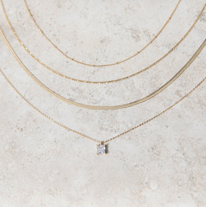 The One Solstice Set by LLUME Jewelry features four gold necklaces displayed on a textured surface: a pendant necklace with a small square gemstone and three stylish gold chains of varying thicknesses, creating an elegant layered look.