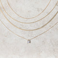 The One Solstice Set by LLUME Jewelry features four gold necklaces displayed on a textured surface: a pendant necklace with a small square gemstone and three stylish gold chains of varying thicknesses, creating an elegant layered look.