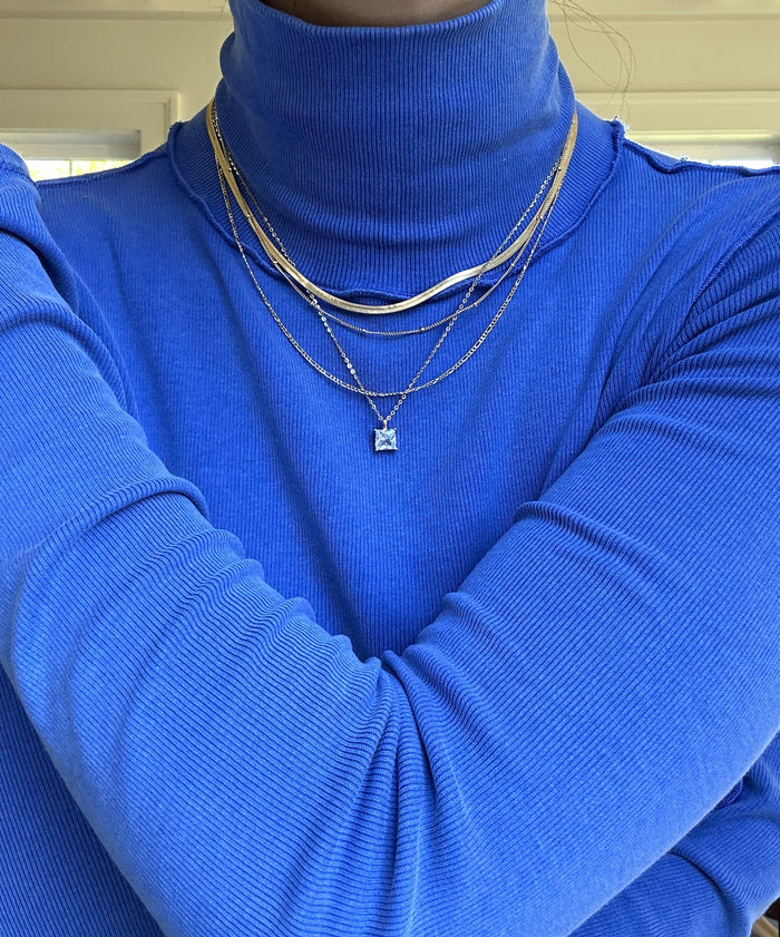 Model wears The One Solstice Set by LLUME Jewelry: a gold chain bundle featuring a square pendant, layered over a royal blue ribbed turtleneck with arms crossed. The softly lit background is illuminated by natural window light.