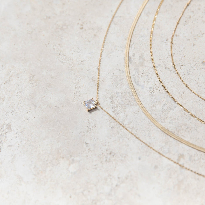 The One Solstice Set by LLUME Jewelry features three delicate gold necklaces arranged in curved lines on a textured stone surface, including a small square-cut clear gemstone pendant for a minimal, elegant layered look.