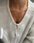 A person in a soft white cardigan wears three delicate gold necklaces from LLUME Jewelry’s Tomato Girl Set, including one with a small red pendant. The focus is on the neck, collarbone, and upper chest.