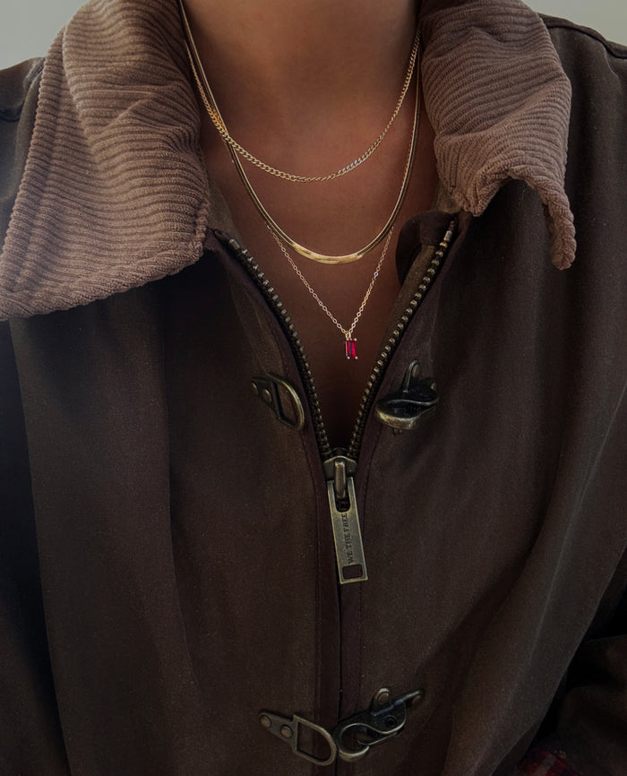 A person wearing a brown corduroy-collared jacket, partially unzipped to reveal stylish layering necklaces—three gold chains, including LLUME Jewelry’s Tomato Girl Set, with two small red pendants.