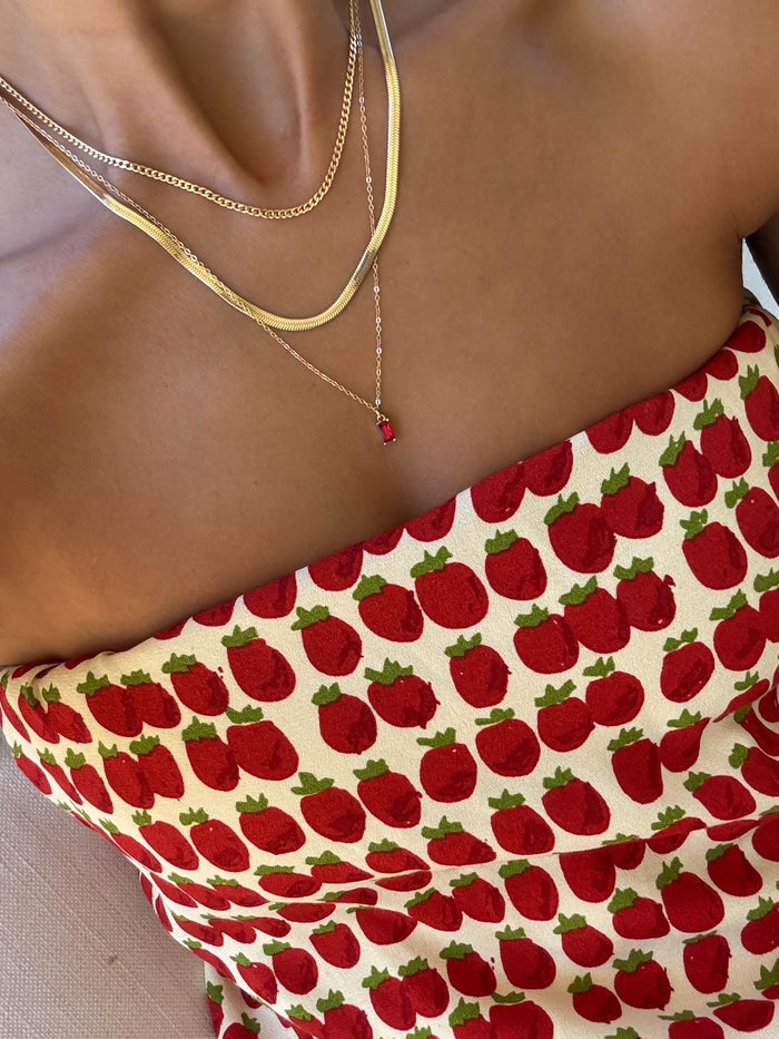 A person is shown from shoulders to upper chest wearing the Tomato Girl Set by LLUME Jewelry—layered necklaces, including one with a small red pendant—paired with a strapless top featuring a red strawberry print.