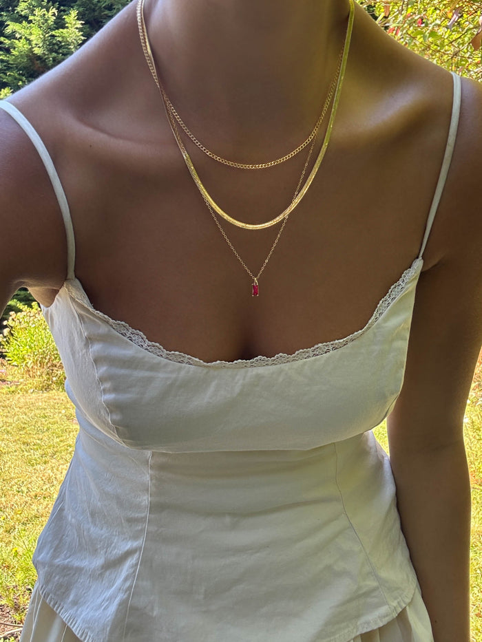 Wearing a white spaghetti strap dress with lace trim and LLUME Jewelry’s Tomato Girl Set—three versatile layering necklaces, one with a pink pendant—a woman stands outdoors in sunlight among lush greenery.