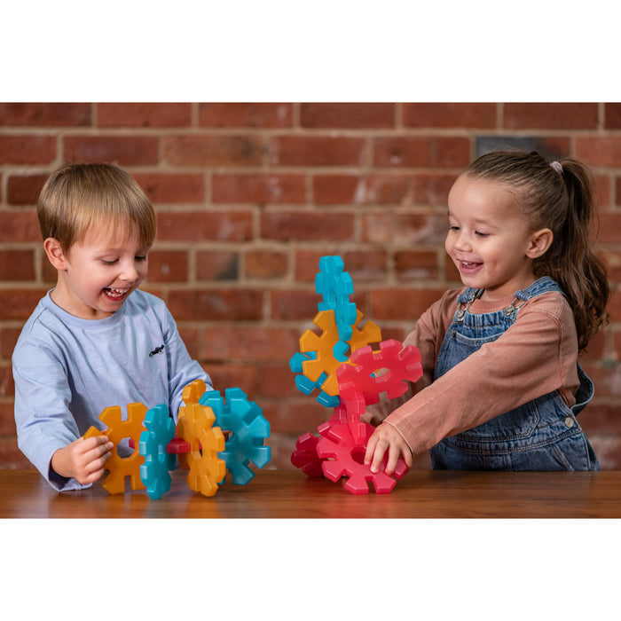 Two children playing with colorful plastic gears on a wooden table against a brick wall.