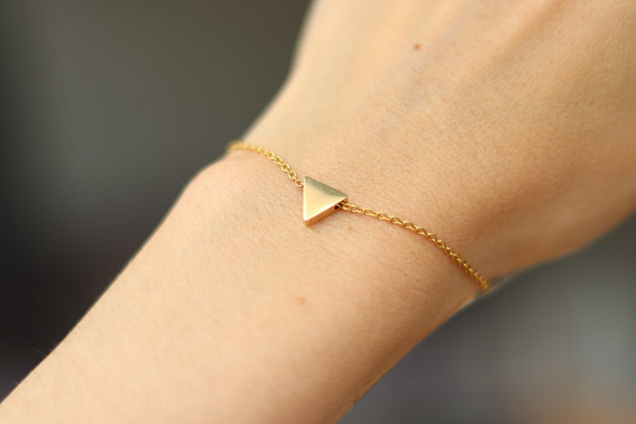 A gold-tone stainless steel chain bracelet with a tiny triangle bead charm, being worn on a person's wrist.