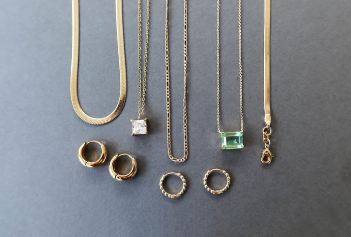 The Vacation Capsule Set by LLUME Jewelry features gold chains with distinctive pendants and chic gold earrings, elegantly displayed on a gray background—your go-to summer jewelry collection.