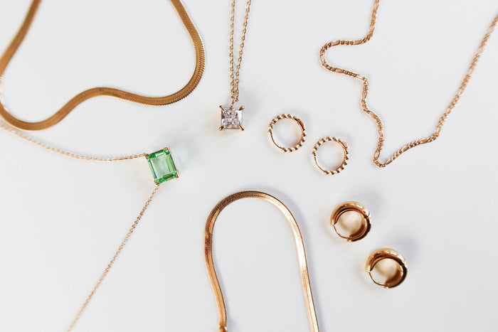 The LLUME Jewelry Vacation Capsule Set features thin gold chain necklaces—one with a green gemstone, one with a square clear stone—and pairs of gold hoop earrings, all arranged on a white surface for a sparkling display.