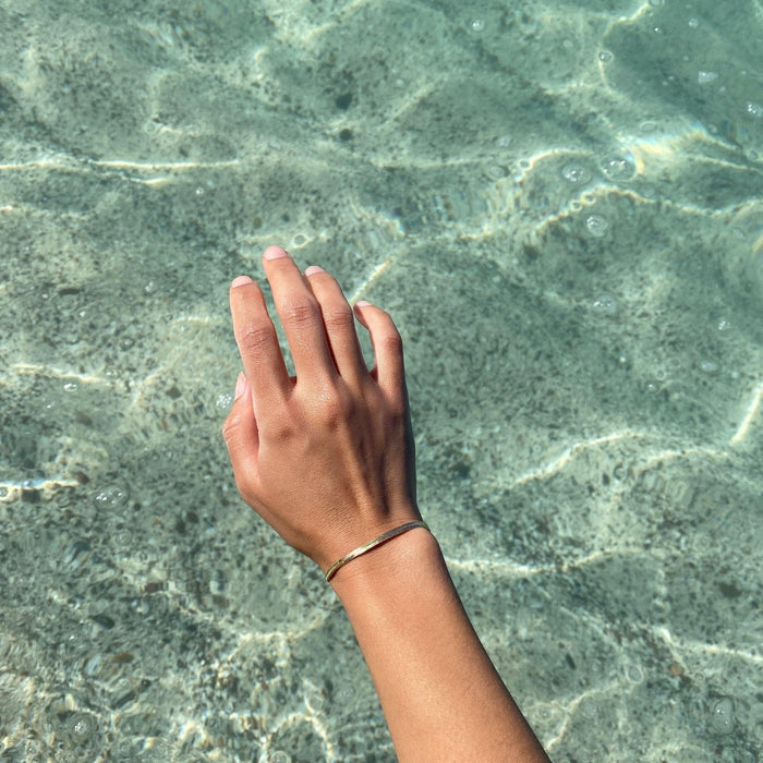 A hand wearing LLUME Jewelry's Vacation Capsule Set is held above sunlit, rippling water, showing how this versatile set can elevate any moment—pair with your favorite pieces for a complete look.
