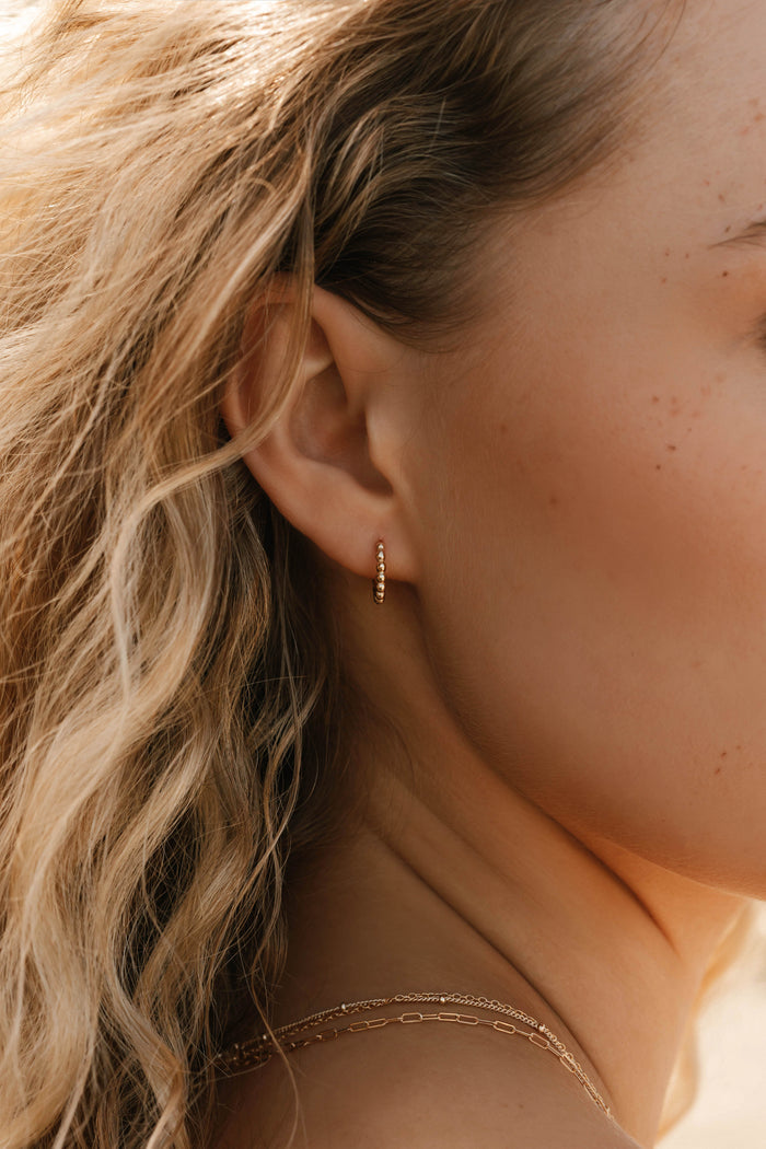A close-up shows wavy blonde hair and an ear adorned with LLUME Jewelry's Vacation Capsule Set—featuring a small gold hoop earring and delicate chain necklaces for an elegant, stackable look.