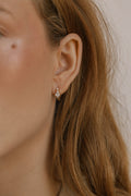 Close-up of a woman with light brown hair wearing a small, elegant gold Vivian Hoop earring by LLUME Jewelry, featuring a clear stone. Smooth skin and a beauty mark are visible near her temple.