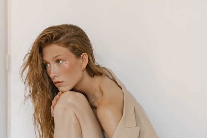 A young woman with long, wavy auburn hair sits against a white background, hugging her knees. She wears a beige blazer draped over her shoulders and LLUME Jewelry's Vivian Hoops, gazing thoughtfully to the side.