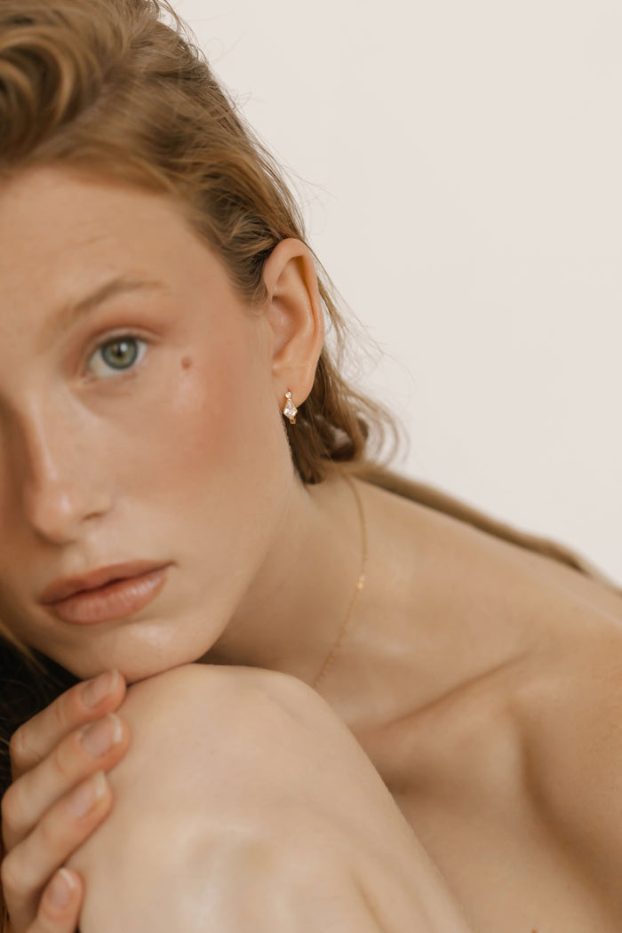 A close-up of a young woman with wavy light brown hair and green eyes rests her chin on her knee, wearing a delicate necklace and LLUME Jewelry's Vivian Hoops, which add an elegant touch against the plain, neutral background.