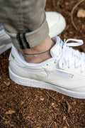 A silver tone chain anklet on a man's ankle, visible above white sneakers.