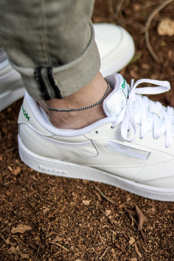 A silver tone chain anklet on a man's ankle, visible above white sneakers.