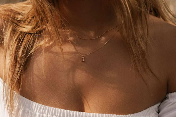 Close-up of a woman with long blonde hair in an off-shoulder white top wearing the LLUME Jewelry Whisper Daily Set—two delicate gold necklaces, including a small vertical Whisper pendant—perfect for everyday wear.