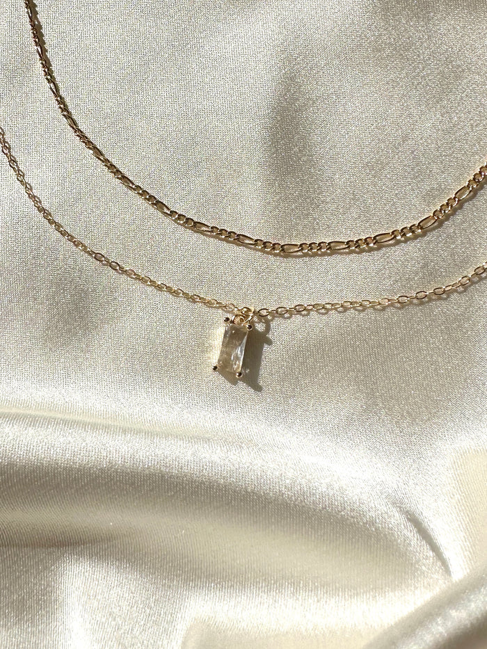 Two delicate gold chains from LLUME Jewelry’s Whisper Daily Set rest on cream satin: one with a rectangular crystal pendant that sparkles, the other a simple, elegant chain—an ideal jewelry set for understated glamour.
