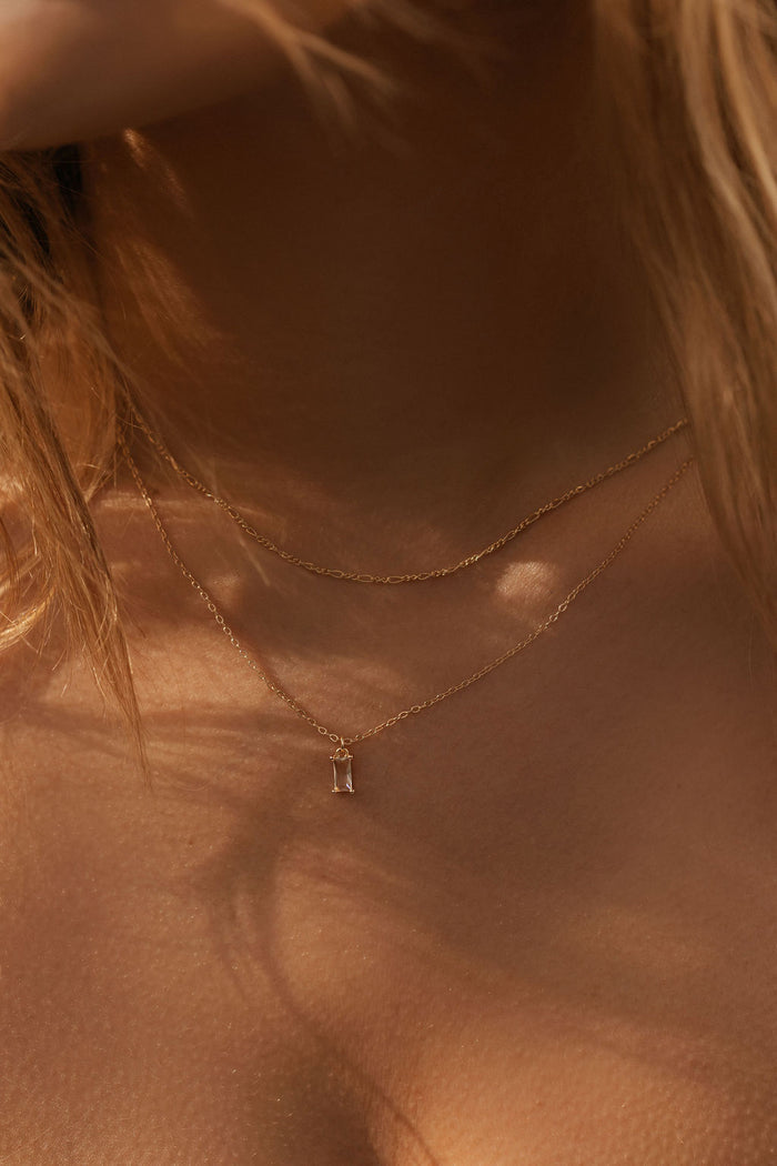 Close-up of a neck wearing LLUME Jewelry's Whisper Daily Set—two delicate gold chain necklaces—in warm, natural light. Strands of light brown hair frame this perfect everyday accessory.