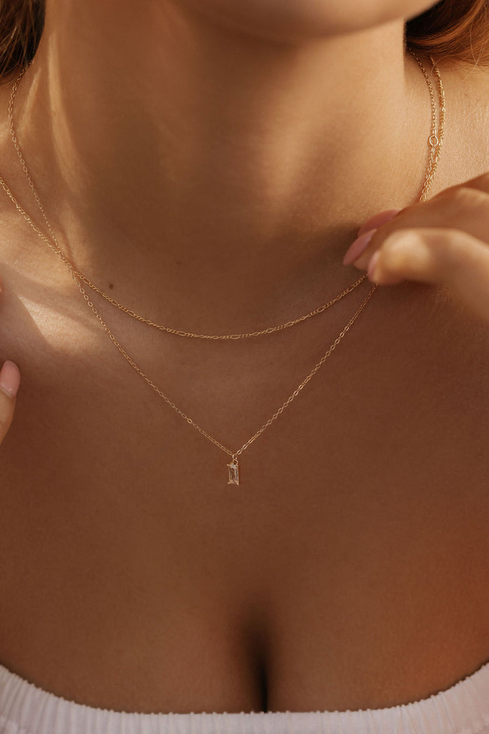 A close-up of a woman wearing the LLUME Jewelry Whisper Daily Set—two delicate gold necklaces, one with a subtle pendant—against bare skin and a white strapless top. Her hands gently touch the chains, highlighting timeless everyday style.