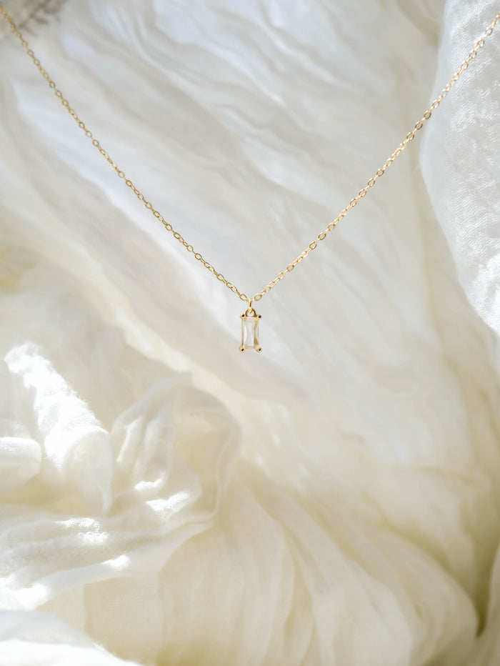 The LLUME Jewelry Whisper Necklace, featuring a delicate gold plated rectangular pendant, is displayed on soft white textured fabric in natural light.