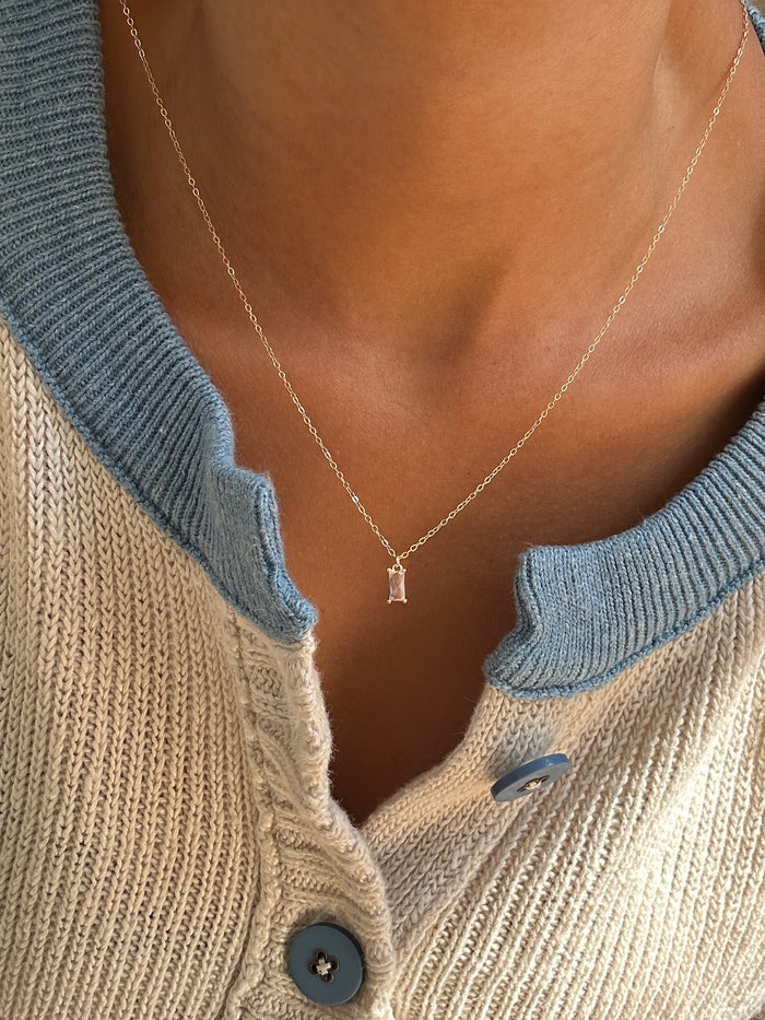 A close-up of a person in a cream knit sweater with blue trim and buttons, styled with LLUME Jewelry’s Whisper Necklace—a gold-plated necklace featuring a delicate small rectangular pendant.