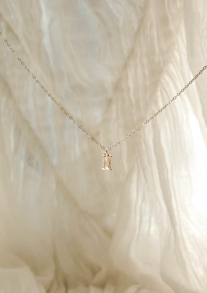 The Whisper Necklace by LLUME Jewelry is a delicate gold-plated piece with a small rectangular pendant, shown against a soft, textured cream-colored fabric background.